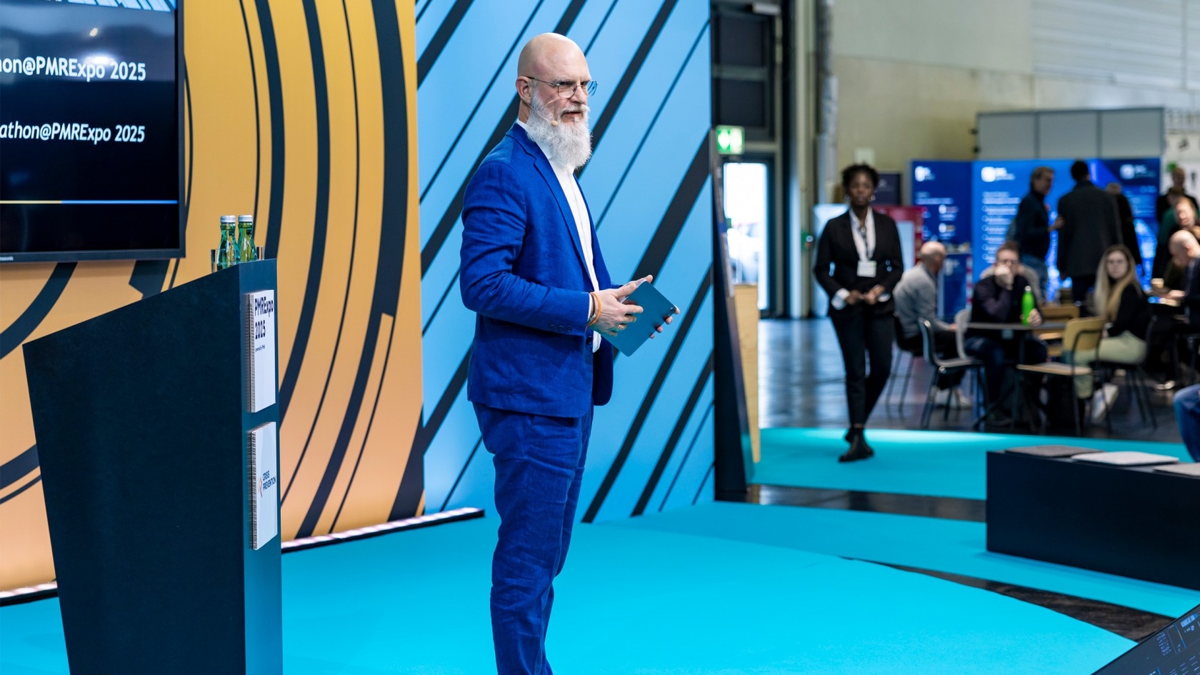 A bearded man in a blue suit confidently presents on stage with a tablet in hand at the bustling PMRExpo 2025, while an engaged audience listens intently in the background.
