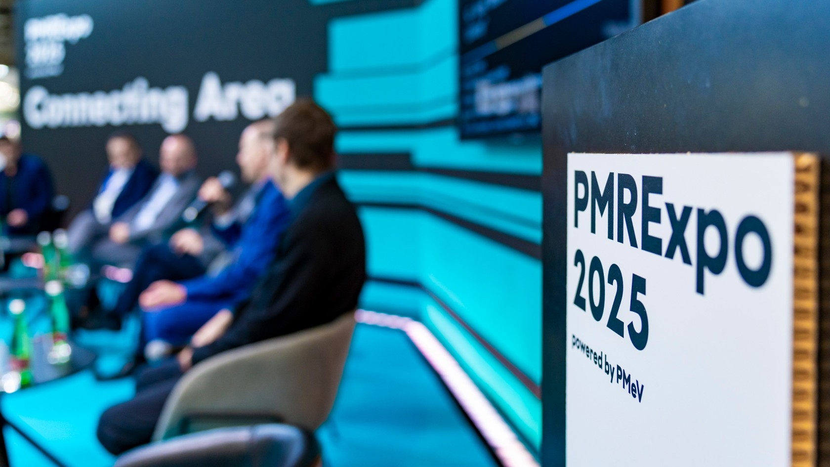 A lively panel discussion unfolds at the PMRExpo 2025, showcasing a diverse group of speakers framed by an eye-catching backdrop adorned with the event's branding.