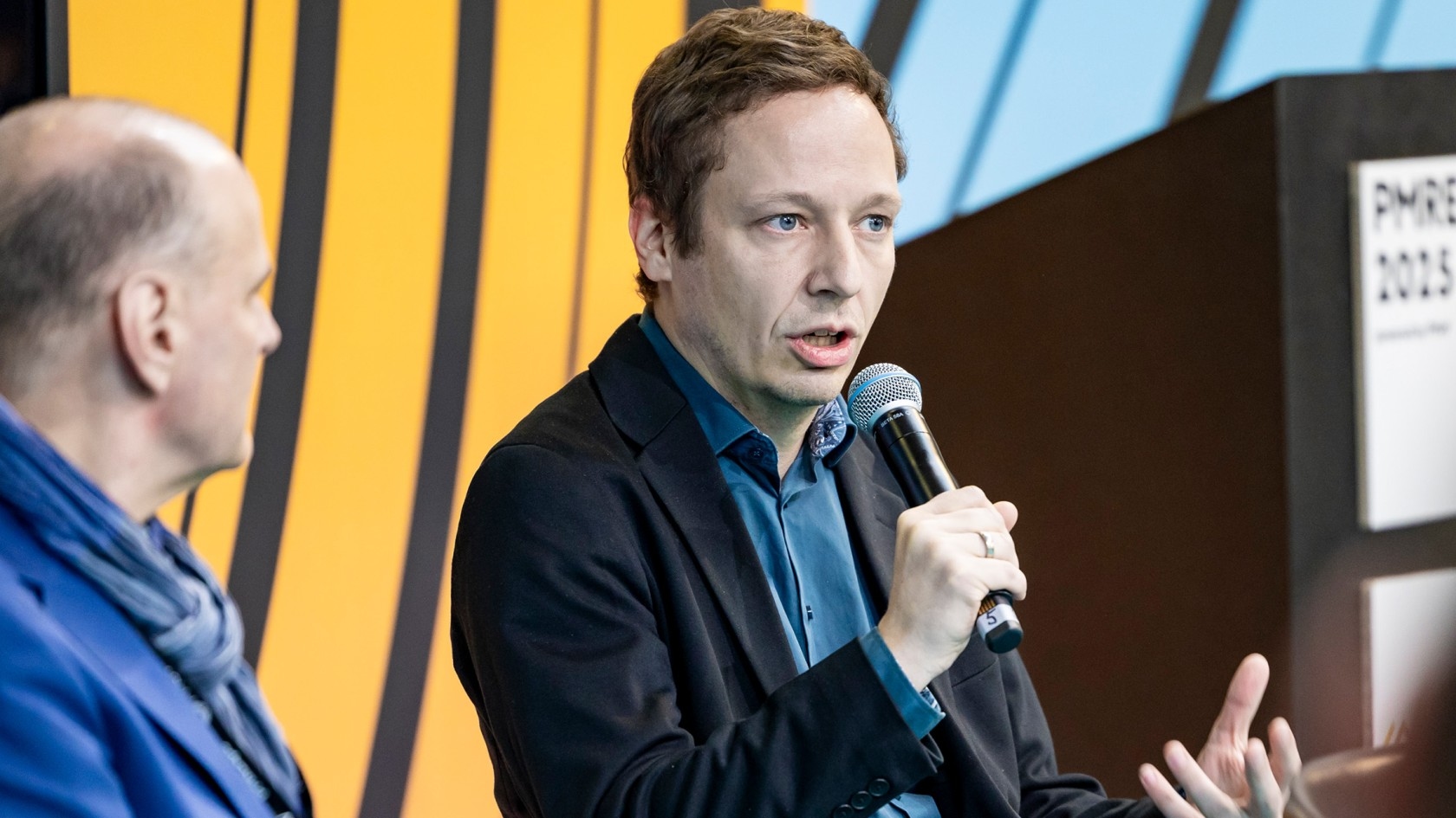 A panelist passionately shares insights into the topic at hand, microphone in hand, while an engaged colleague listens attentively in a formal conference environment.