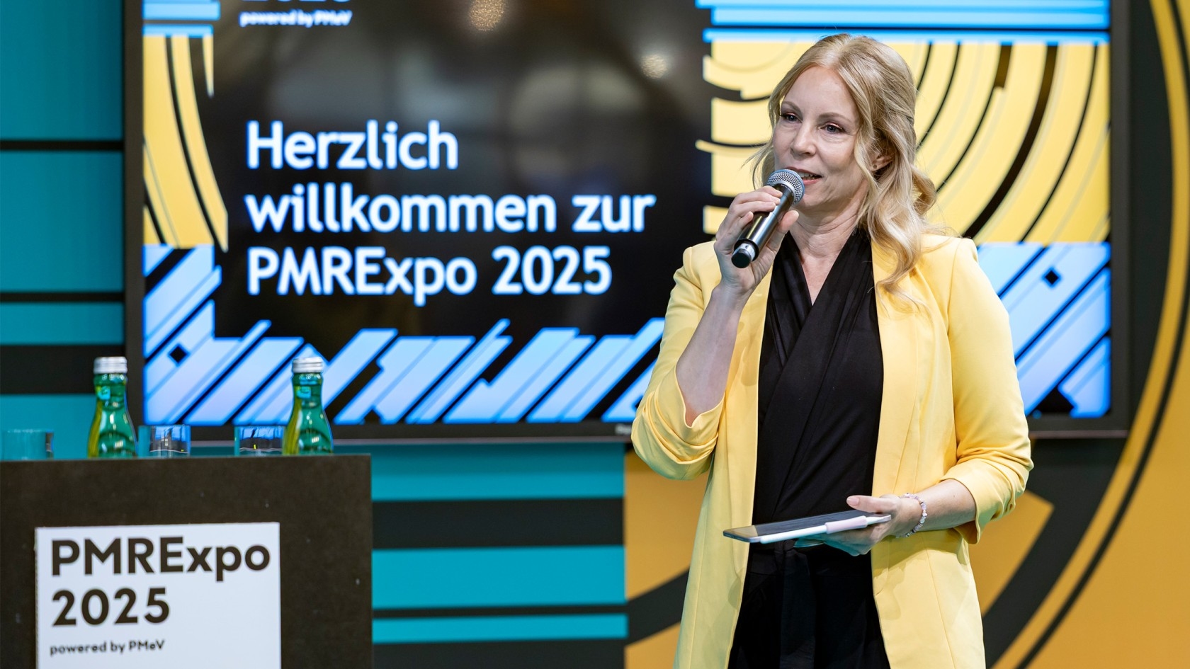 A confident woman in a vibrant yellow blazer addresses the audience from the stage, holding a microphone as a banner behind her proudly showcases "Welcome to PMRexpo 2025."
