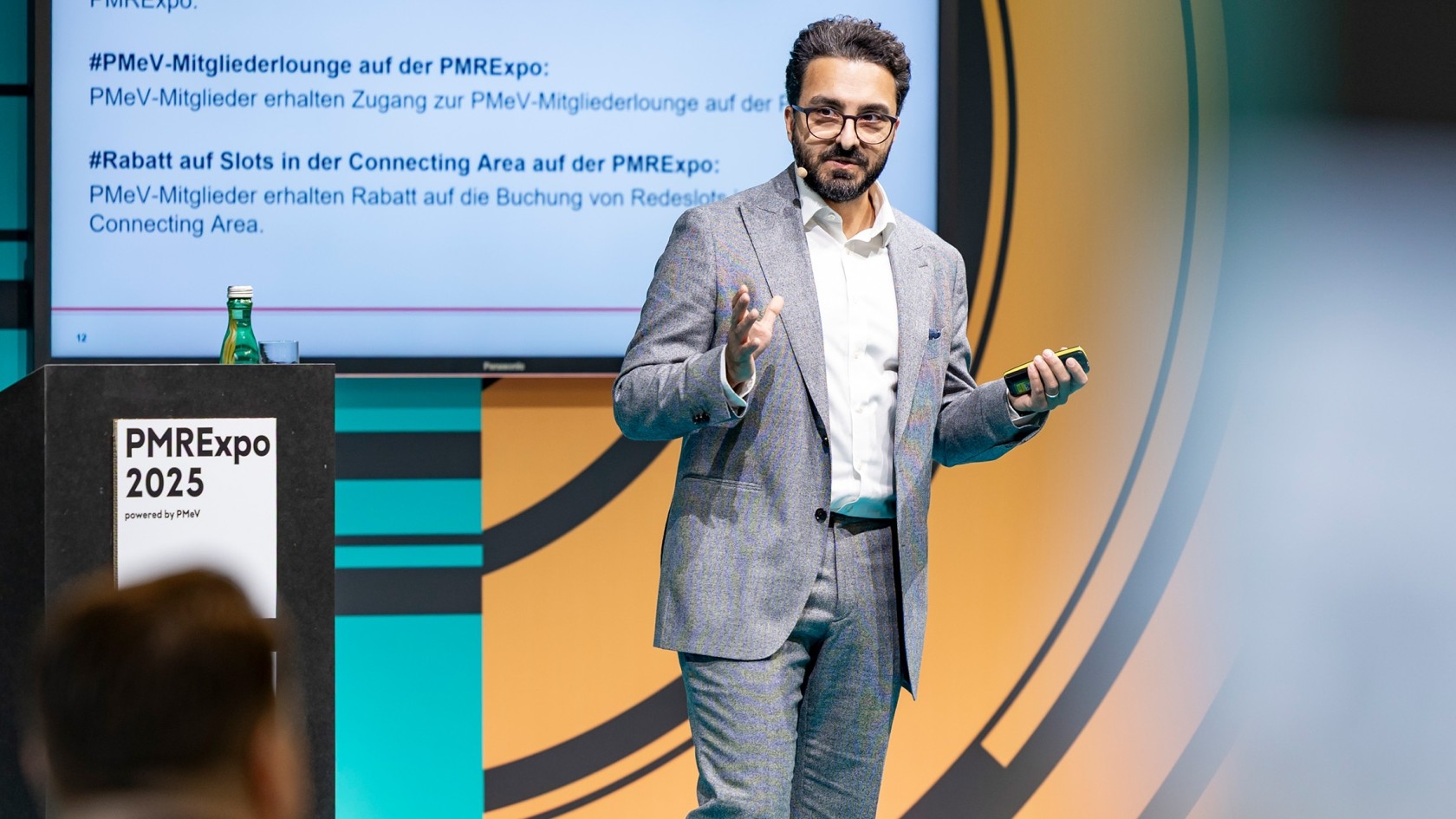 In a sleek conference hall, a presenter in a stylish gray suit animatedly gestures as they share insights on PMRExpo 2025, with a vibrant slide showcasing the event's highlights prominently displayed behind them.