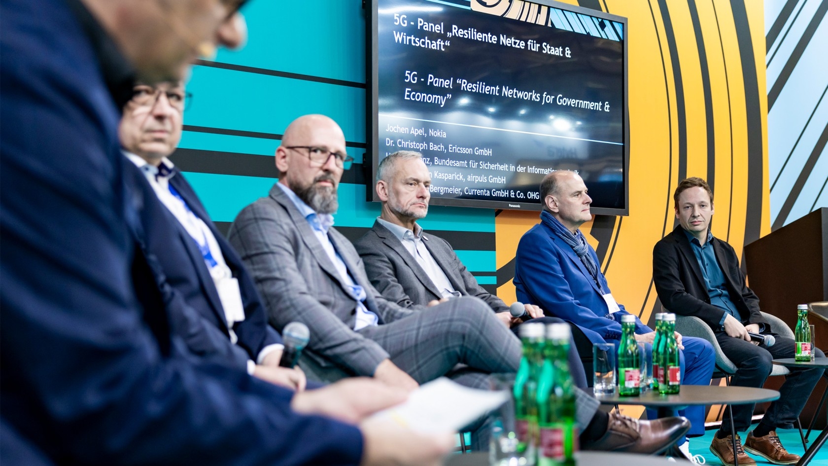 A dynamic panel discussion on resilient networks for government and economy unfolds, showcasing five insightful speakers engaging in dialogue, with refreshments set on tables and a sleek digital presentation screen enhancing the experience.