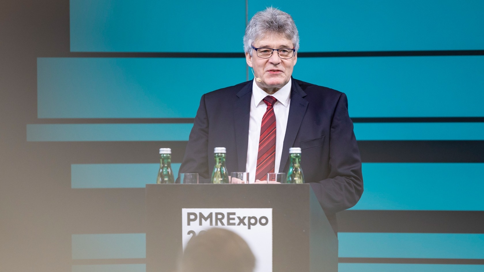 A polished speaker stands confidently at a podium adorned with water bottles, engaging the audience during the prominent PMRexpo 2023 event set against an elegant blue backdrop.