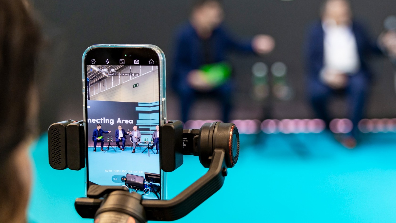 A smartphone mounted on a stabilizer records a panel discussion on stage, where speakers engage in dialogue against a captivating backdrop emblazoned with "Connecting Area."