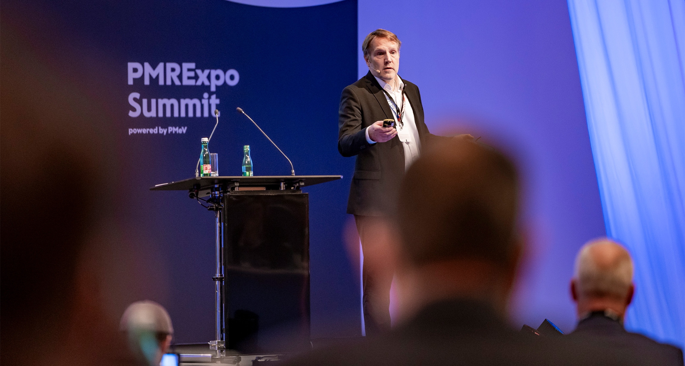 A speaker confidently addresses the audience from a podium at the PMRExpo Summit, engaging with the attentive crowd in the foreground while water bottles are neatly placed on the table beside them.