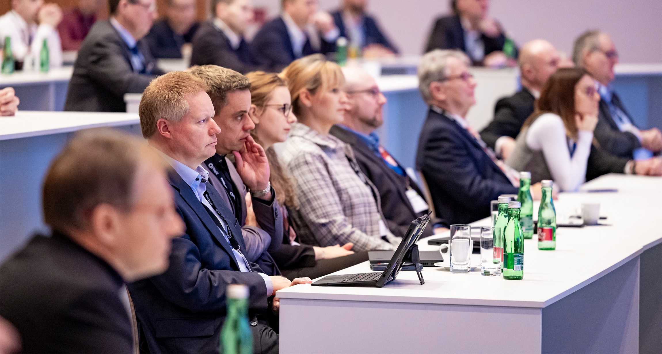 Attendees engage with rapt attention at a vibrant workshop, seated around modern tables adorned with refreshing water bottles and glasses, as they face an inspiring speaker.