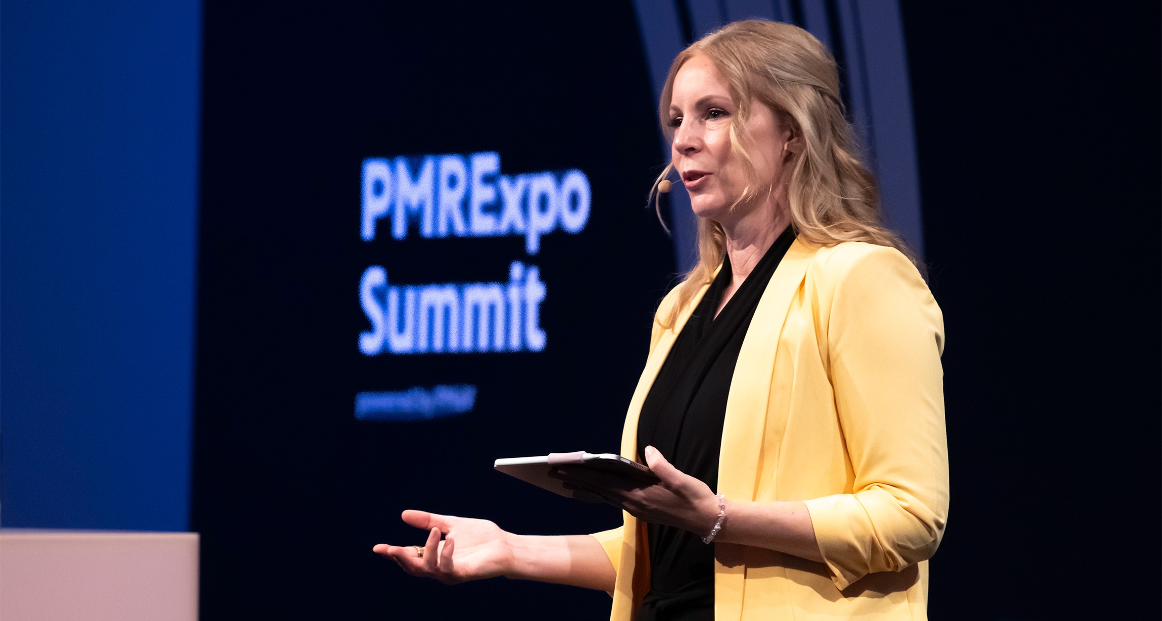 A confident woman in a vibrant yellow blazer addresses the audience at the PMRExpo Summit, energetically gesturing with a tablet in hand to emphasize her points.