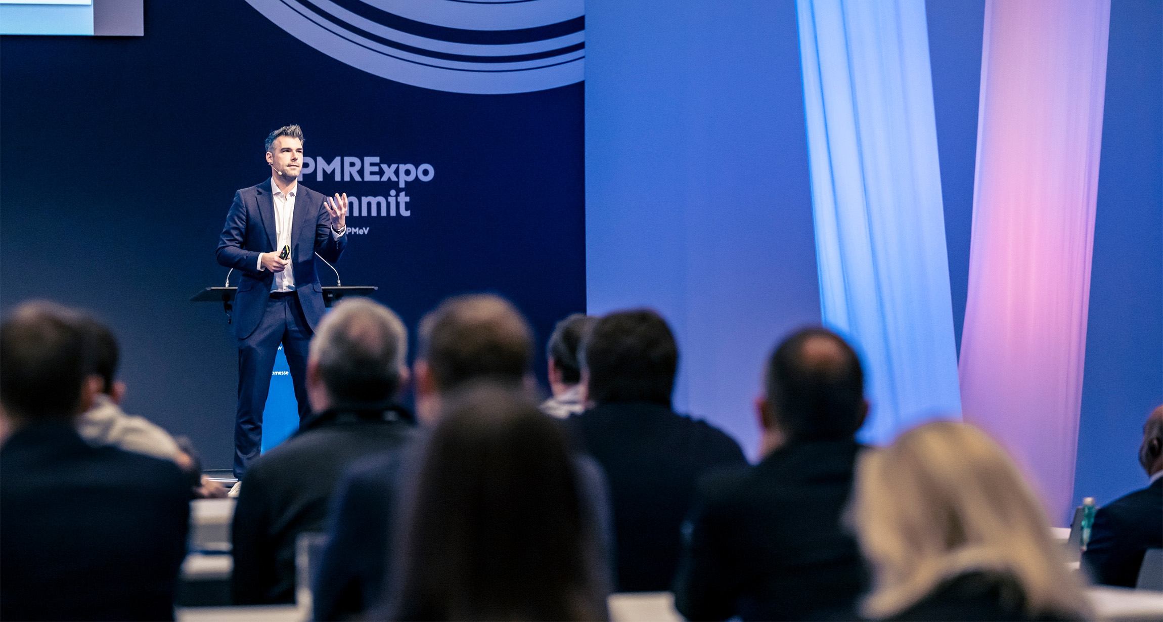 A dynamic speaker stands confidently on stage at a vibrant conference, engaging an attentive audience seated at elegantly arranged tables, while colorful lighting casts a lively glow on the backdrop.
