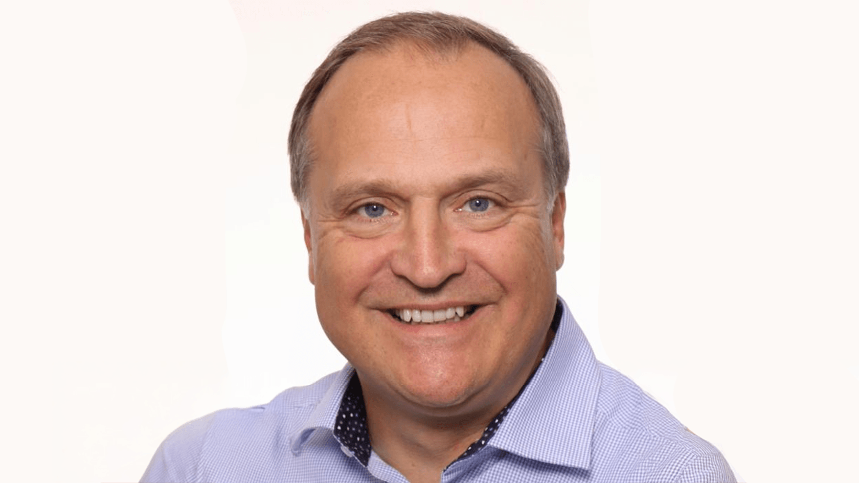 A smiling man with short hair, dressed in a light blue shirt, stands confidently against a simple white backdrop, embodying the innovative spirit of PMRExpo as he represents his company's cutting-edge solutions and products.