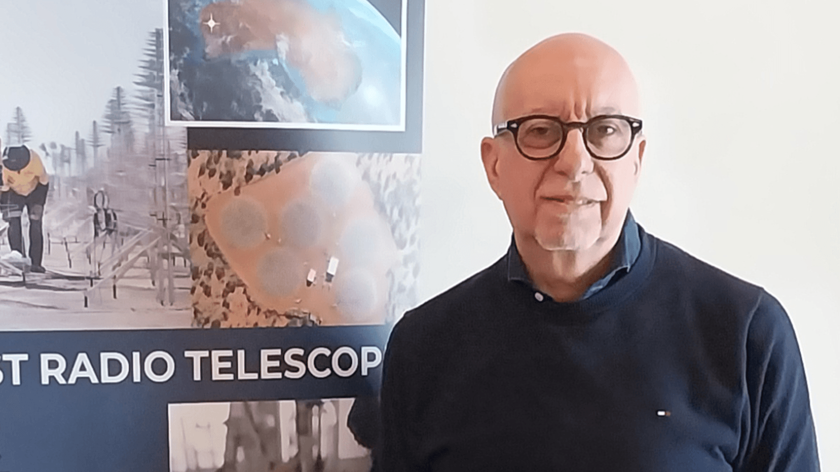 A man with glasses stands in front of a poster showcasing radio telescopes and advancements in astronomical research, reflecting Sirio Antenne SRL's commitment to innovation at PMRExpo.