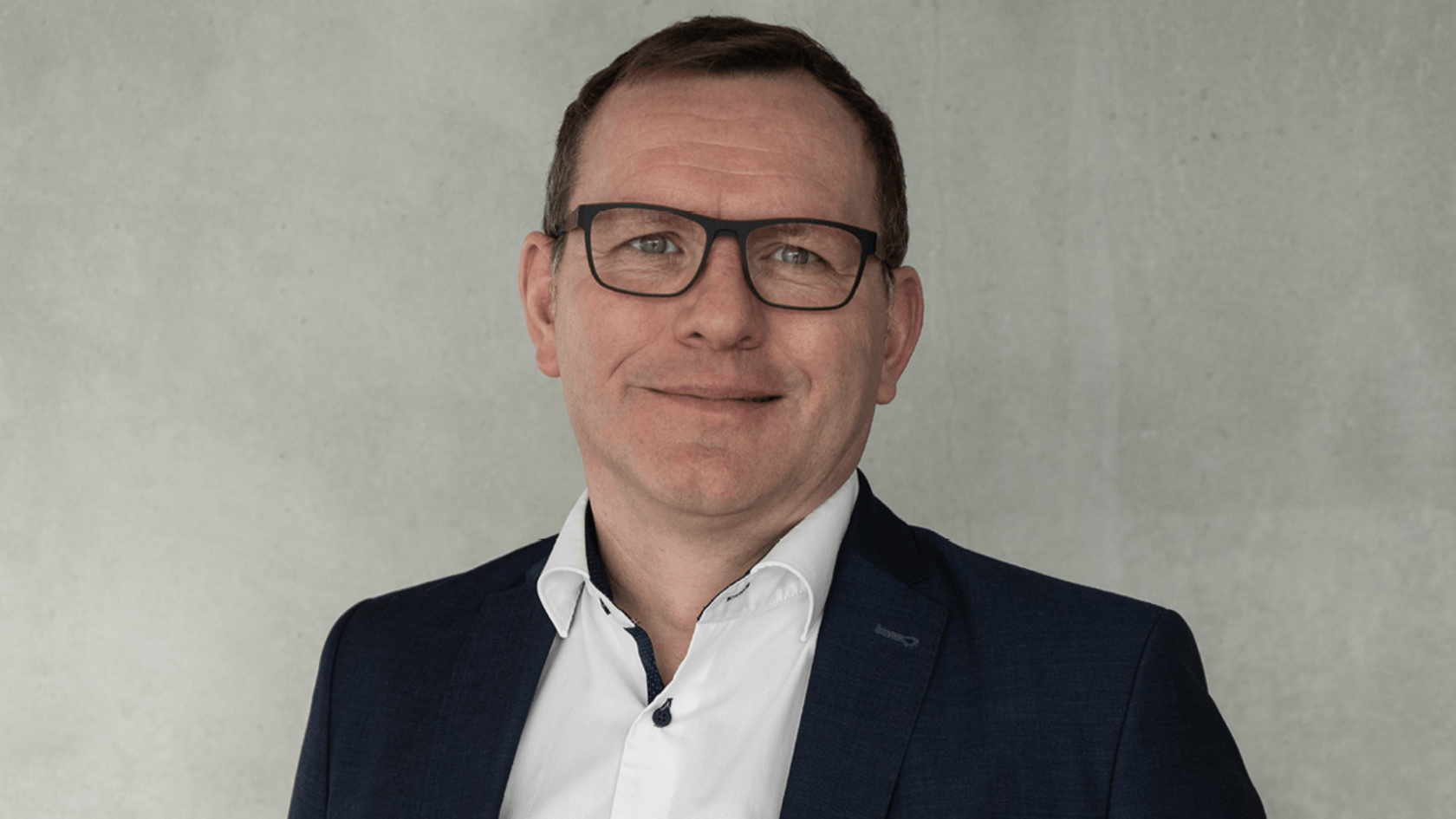 A smiling man with glasses, dressed in a dark suit jacket over a crisp white shirt, stands confidently against a backdrop of sleek gray concrete, embodying the professionalism and innovation that MSA Deutschland GmbH aims to showcase at PMRExpo.