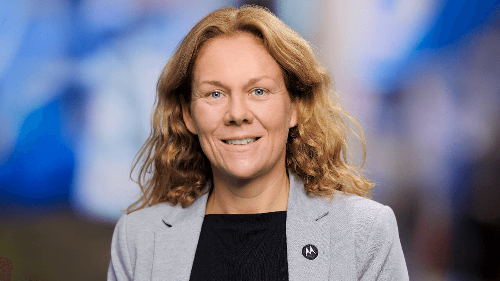 A smiling woman with curly hair, dressed in a grey blazer over a black shirt, stands confidently in front of a softly blurred blue backdrop, embodying the innovative spirit of Motorola Solutions at PMRExpo.