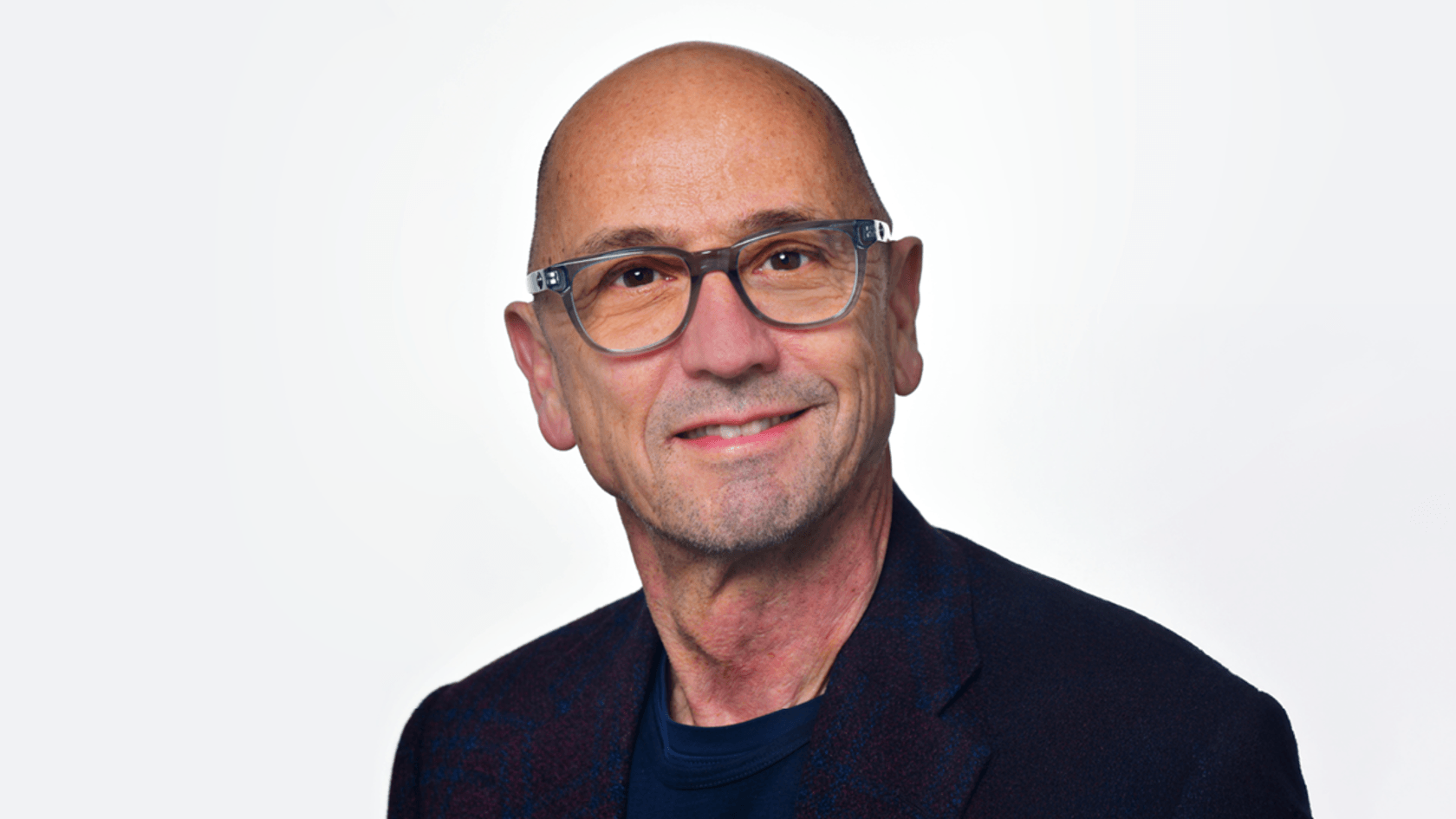 A man with a bald head and glasses smiles confidently in a dark blazer over a navy shirt, embodying the spirit of collaboration and innovation that defines the PMRExpo event, set against a light background.