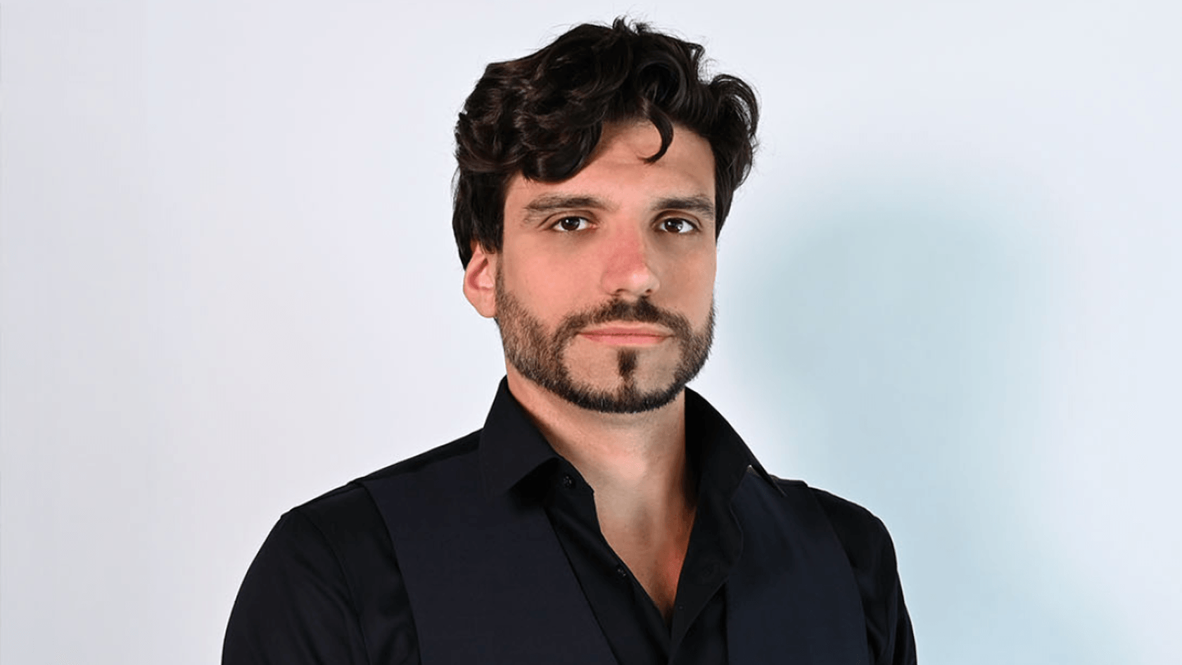 A man with curly dark hair and a beard, dressed in a sleek black vest and shirt, confidently poses against a light backdrop, embodying the innovative spirit of PMRExpo as he represents sureVIVE SA and shares insights on the company's groundbreaking solutions and products.