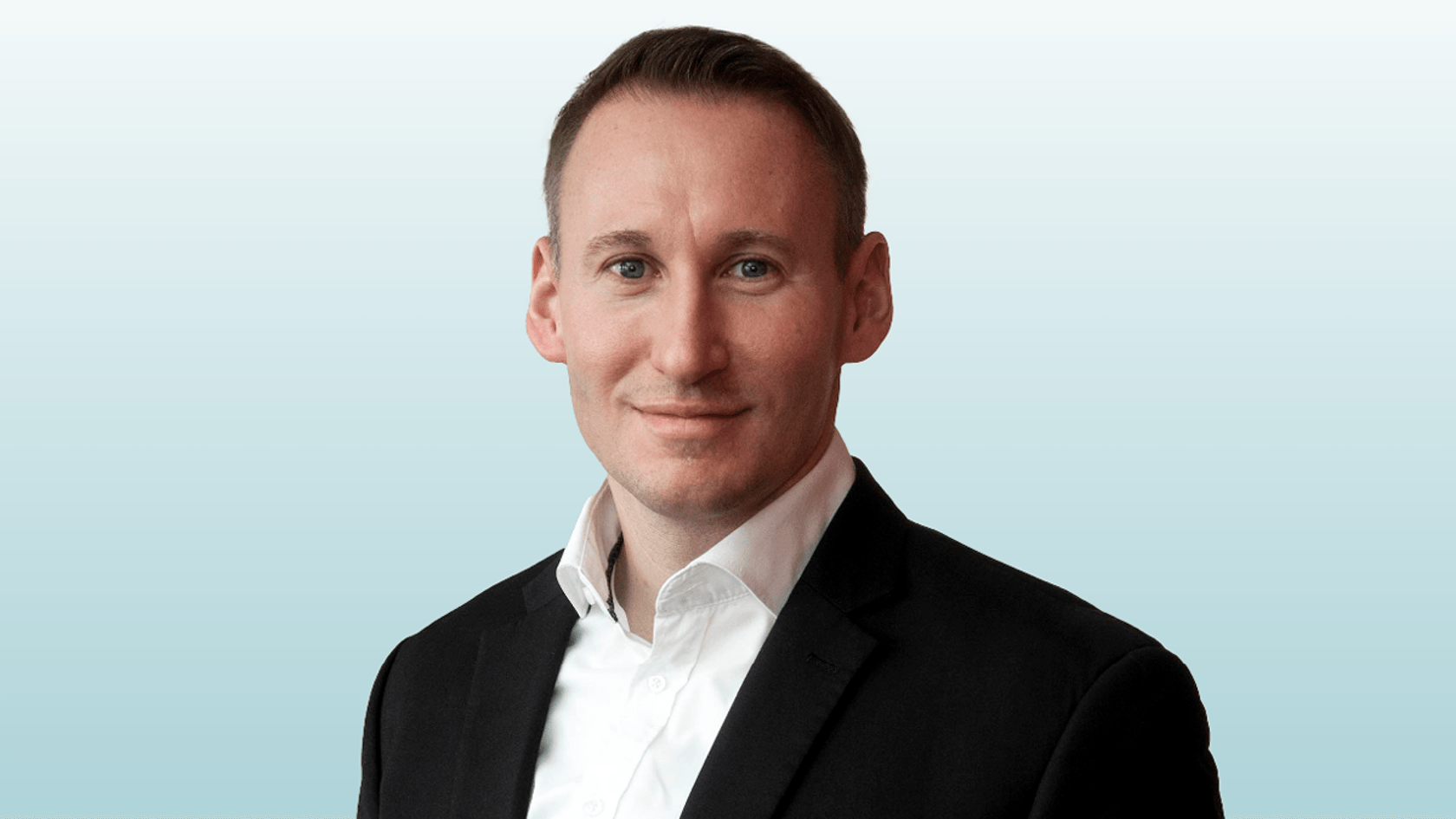 A confident Holger Klaus, dressed in a sharp black suit with a crisp white shirt, smiles as he stands against a light gradient background, embodying the innovation and expertise showcased at PMRExpo.
