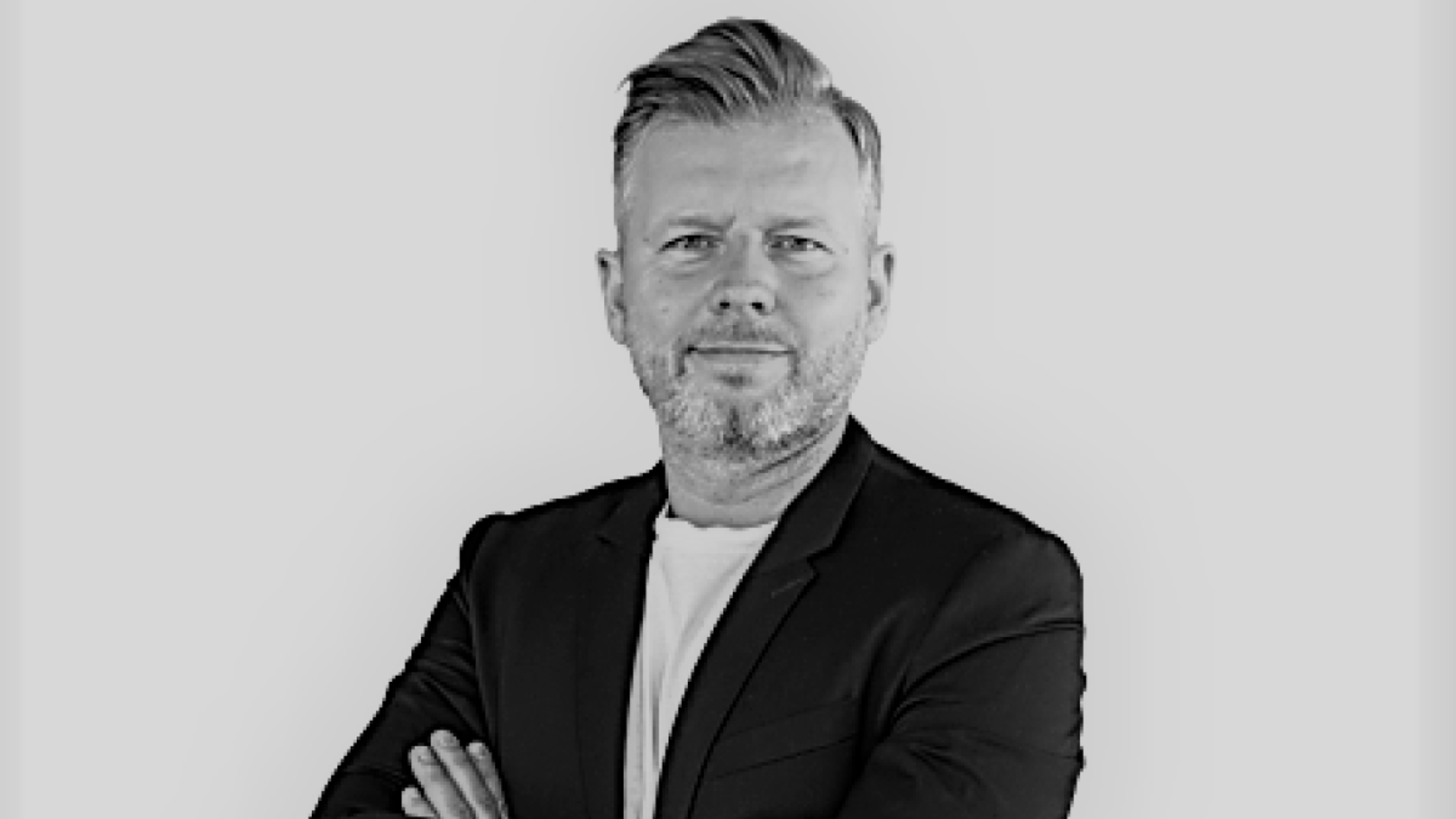 A confident man with short hair and a well-groomed beard stands with arms crossed in a black blazer over a white shirt, against a light background, embodying the expertise and leadership of ASC Technologies AG as he prepares to engage with industry professionals at PMRExpo.