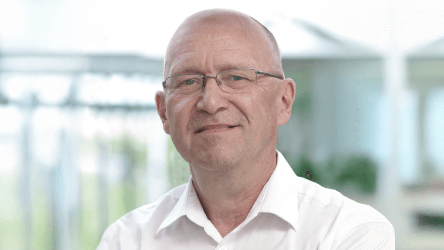 A middle-aged man with glasses, wearing a white shirt, stands confidently and smiles warmly amidst a bright, modern exhibition space filled with greenery, poised to showcase innovative products and engage with attendees at PMRExpo.