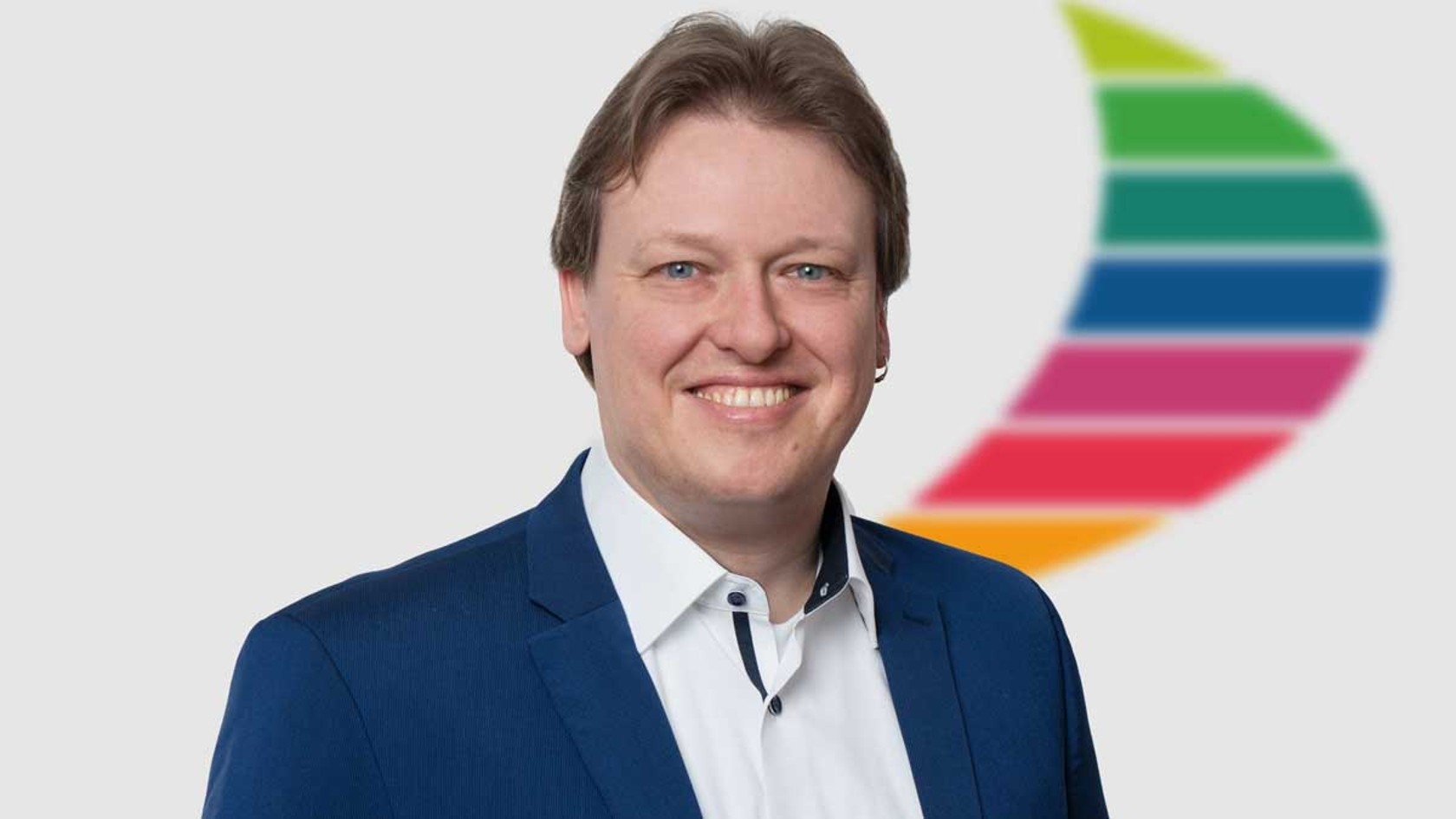 A smiling man in a blue suit stands confidently against a light gray backdrop, with a colorful logo partially visible next to him, representing his role as an Executive Officer at the pivotal PMRExpo, where public safety organizations and IT solutions converge.
