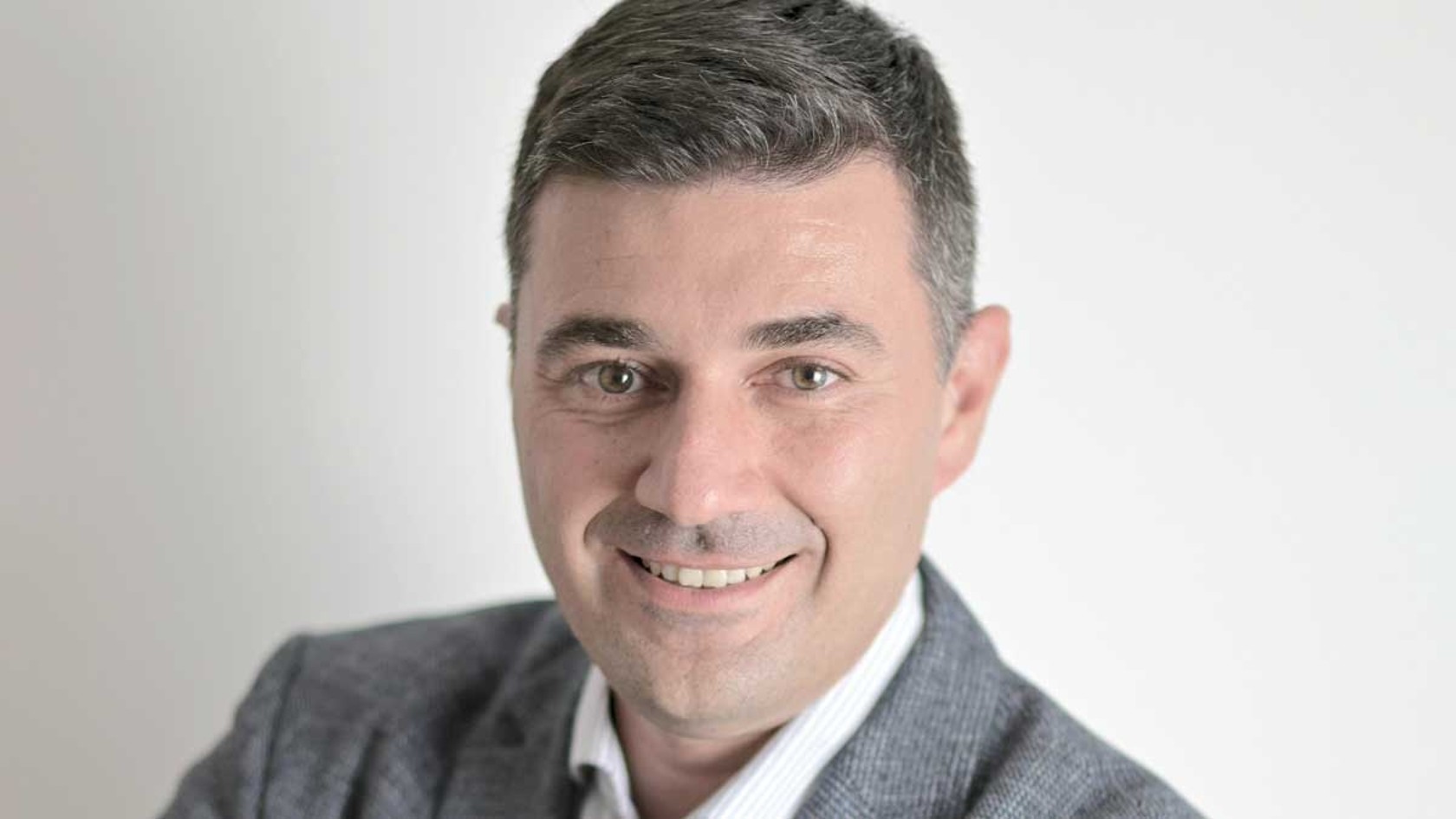 A smiling man with short dark hair and a light gray blazer stands confidently against a minimalist backdrop, embodying the innovative spirit of Schnoor at PMRExpo, where he represents their commitment to quality and collaboration in critical communications.