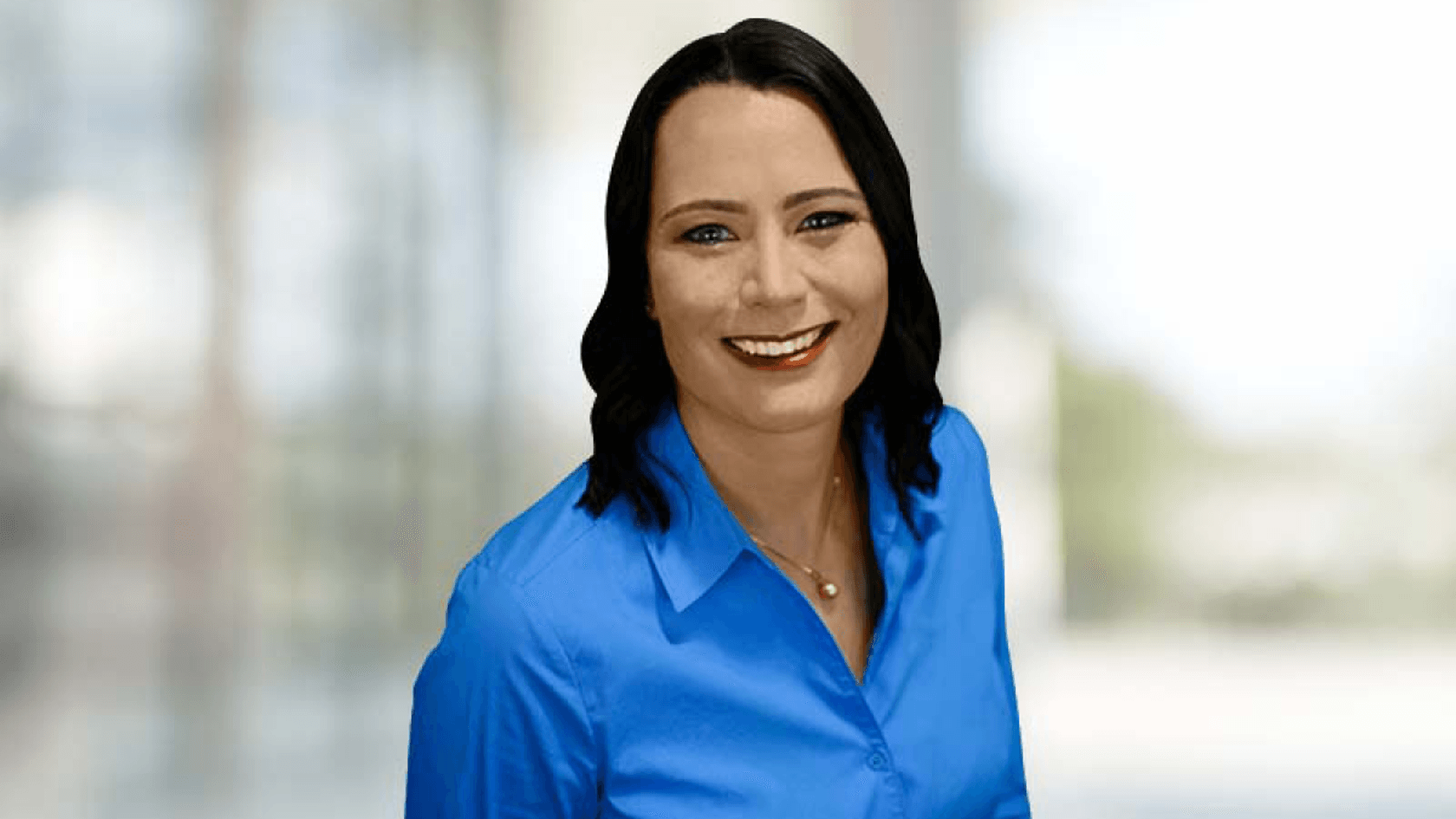 A confident Tina Mucha, Global Marketing Manager of Omnitronics Pty Ltd, smiles as she stands in front of a vibrant, dynamic backdrop, showcasing her enthusiasm while dressed in a professional blue shirt that reflects her company’s innovative spirit.