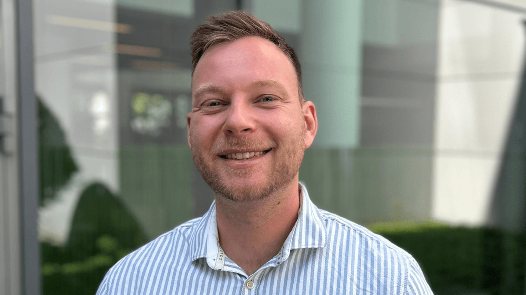 A smiling man with short, light hair wearing a striped shirt stands outdoors at PMRExpo, showcasing innovative solutions for security-critical communications against a backdrop of modern buildings and lush greenery.