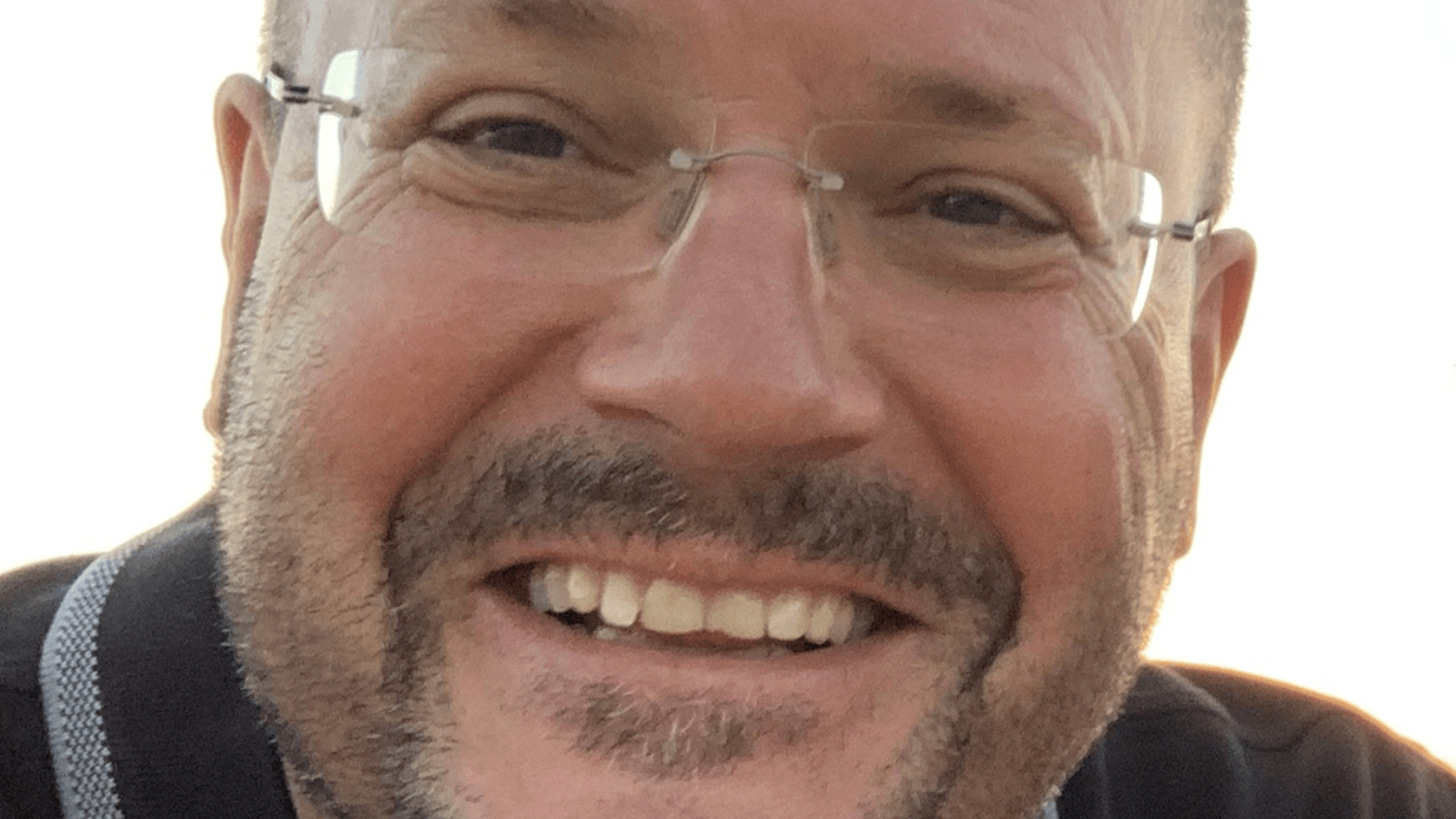 A smiling man with glasses and short hair, sporting a beard and wearing a dark shirt, stands close-up against a light background, embodying the approachable and innovative spirit of NoraTec GmbH as he prepares to engage with decision makers at PMRExpo.