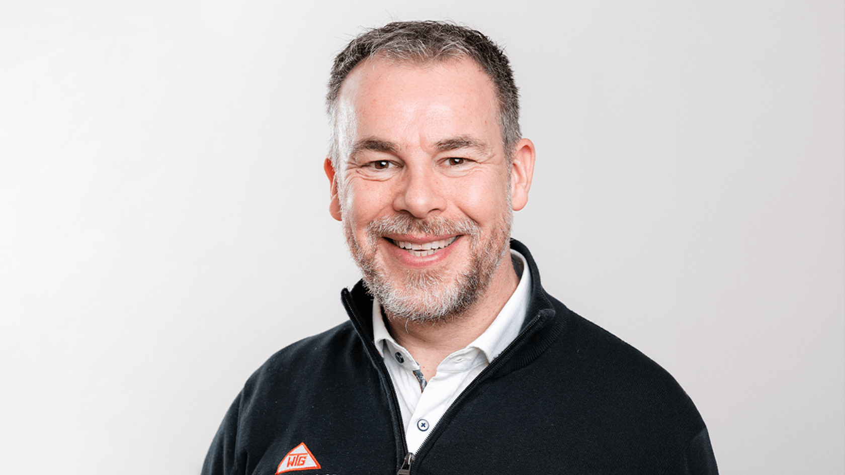 A smiling man with a beard, dressed in a sleek black zip-up jacket over a collared shirt, stands confidently against a light backdrop as he prepares to discuss his expectations and innovative solutions for companies at PMRExpo.