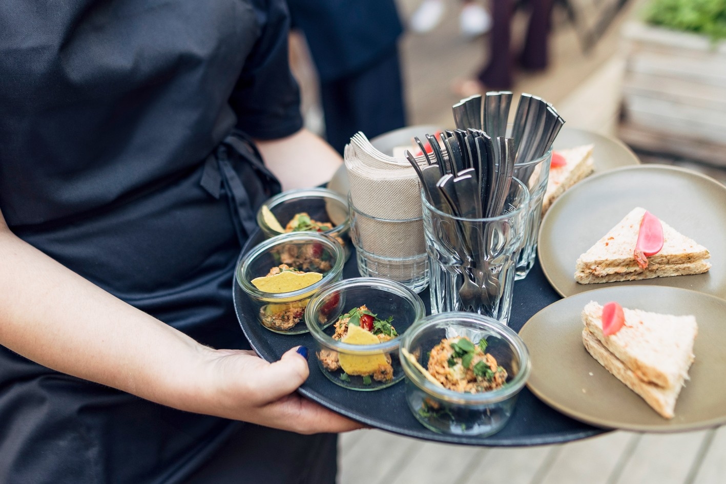 Stand catering Server holding tray with small glasses of appetizers, cutlery, and plates with triangular sandwiches topped with pink flower petals.