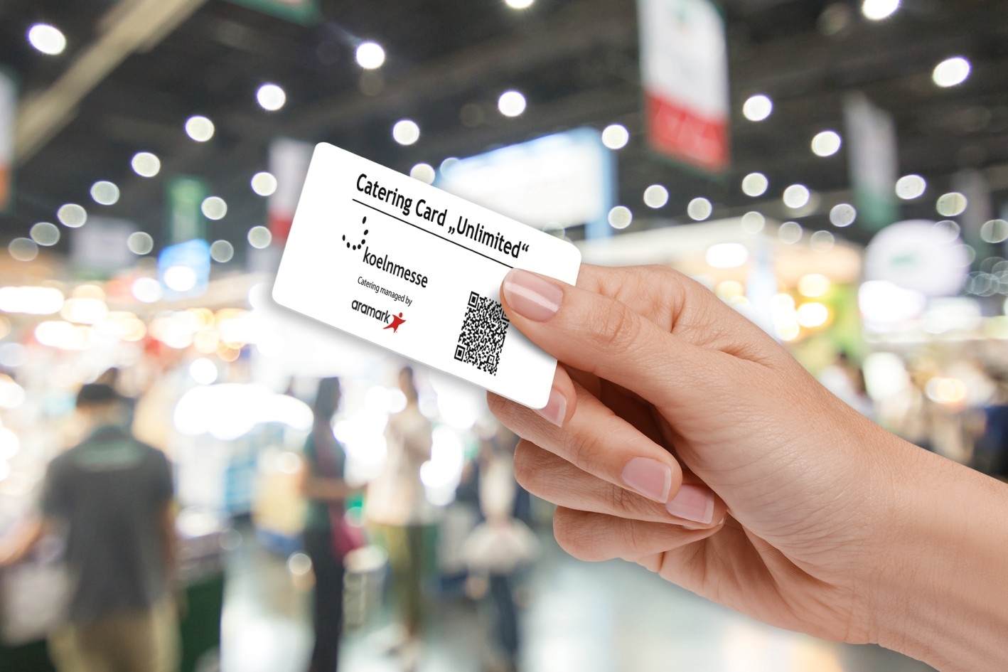 Catering Card Hand holding a white card with the text 'Catering Card Unlimited', a QR code, and logos against a blurred trade fair background.