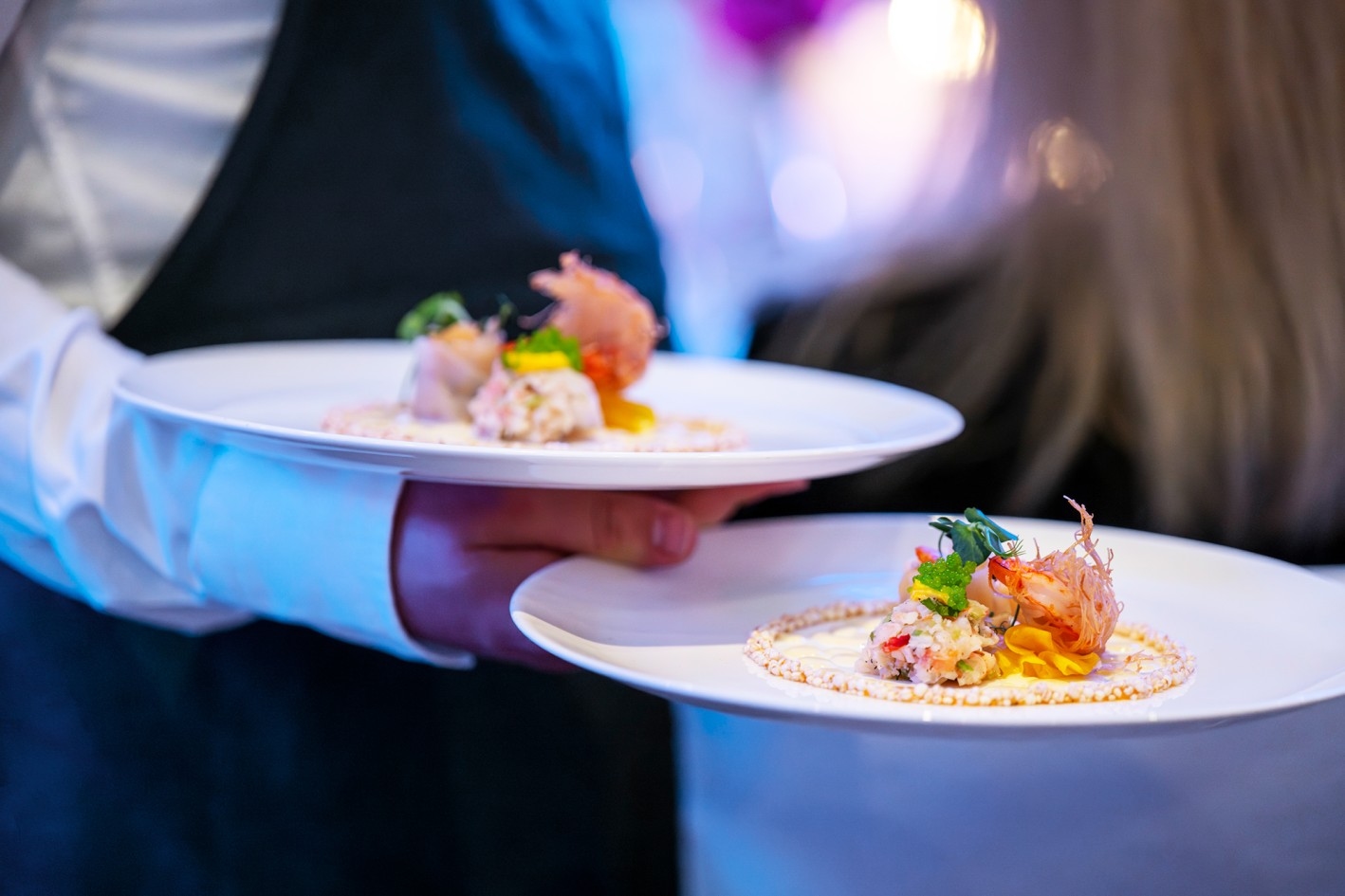 Conference catering Waiter holding two white plates with artfully arranged appetizers including shrimp and herbs.