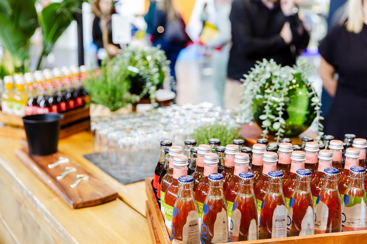 Events accompanying the trade fair Multiple rows of colorful glass beverage bottles on a wooden table, blurred people and green plants in the background.
