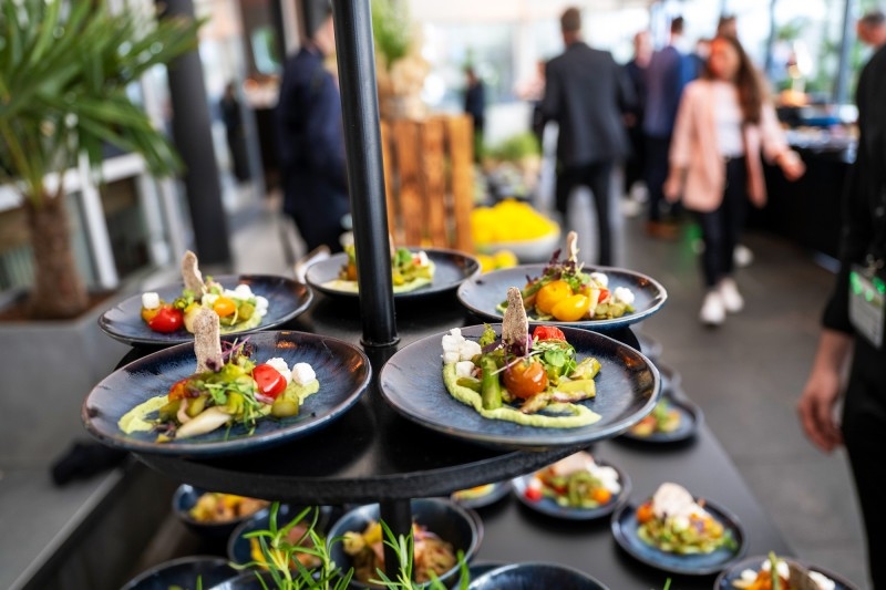 Catering Multiple black plates with artfully arranged appetizers on a multi-tiered serving stand in a busy indoor setting.