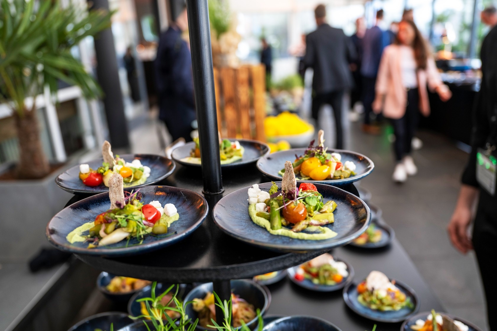 Multiple black plates with artfully arranged appetizers on a multi-tiered serving stand in a busy indoor setting.