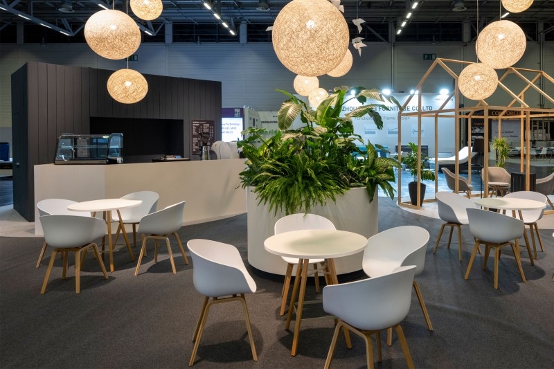 VIP Lounge Several round tables with white chairs surround a large planter with green plants in a modern indoor space with hanging round lights.