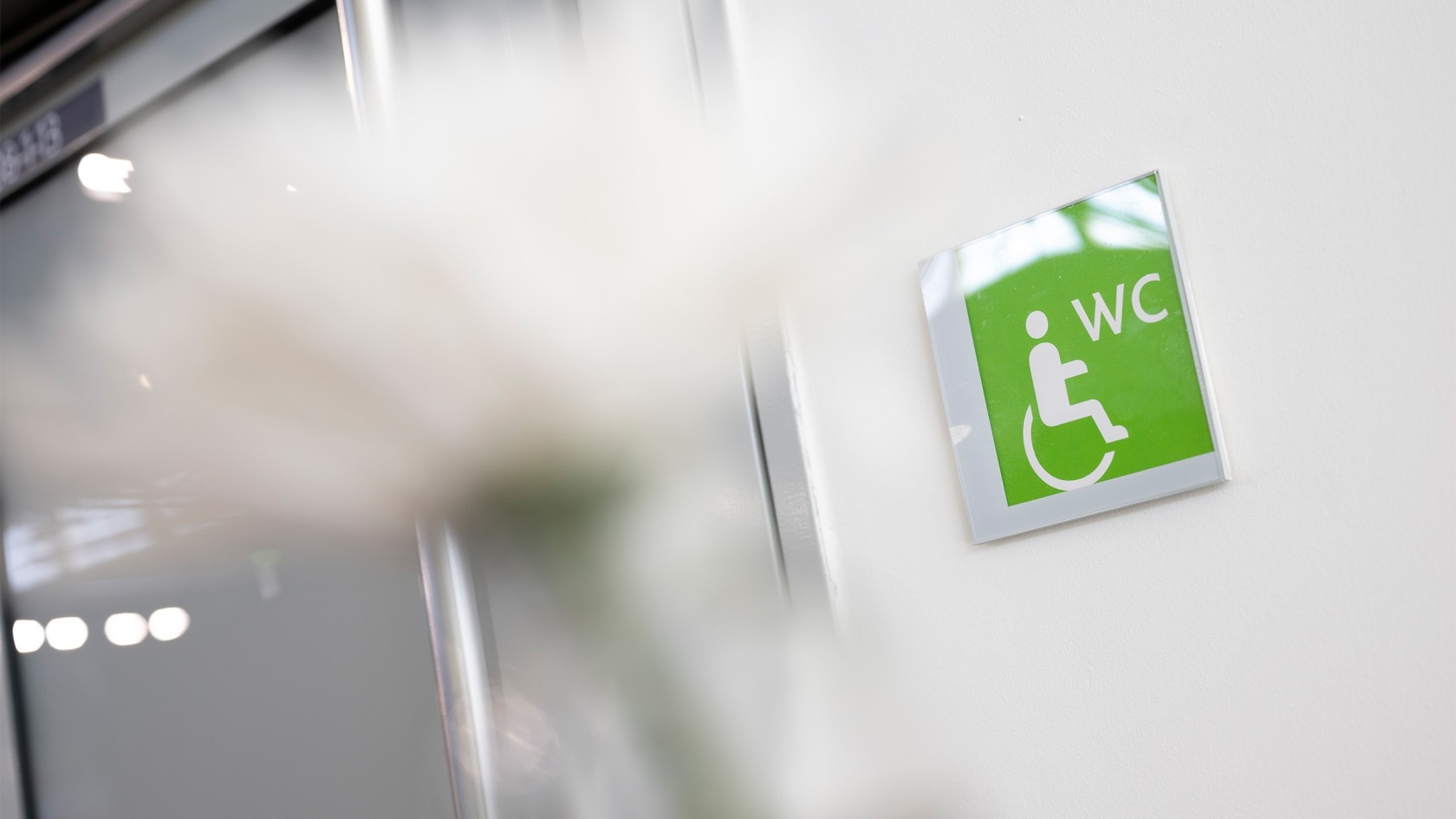 Grünes Schild mit weißem Rollstuhlsymbol und den Buchstaben 'WC' an einer weißen Wand.
